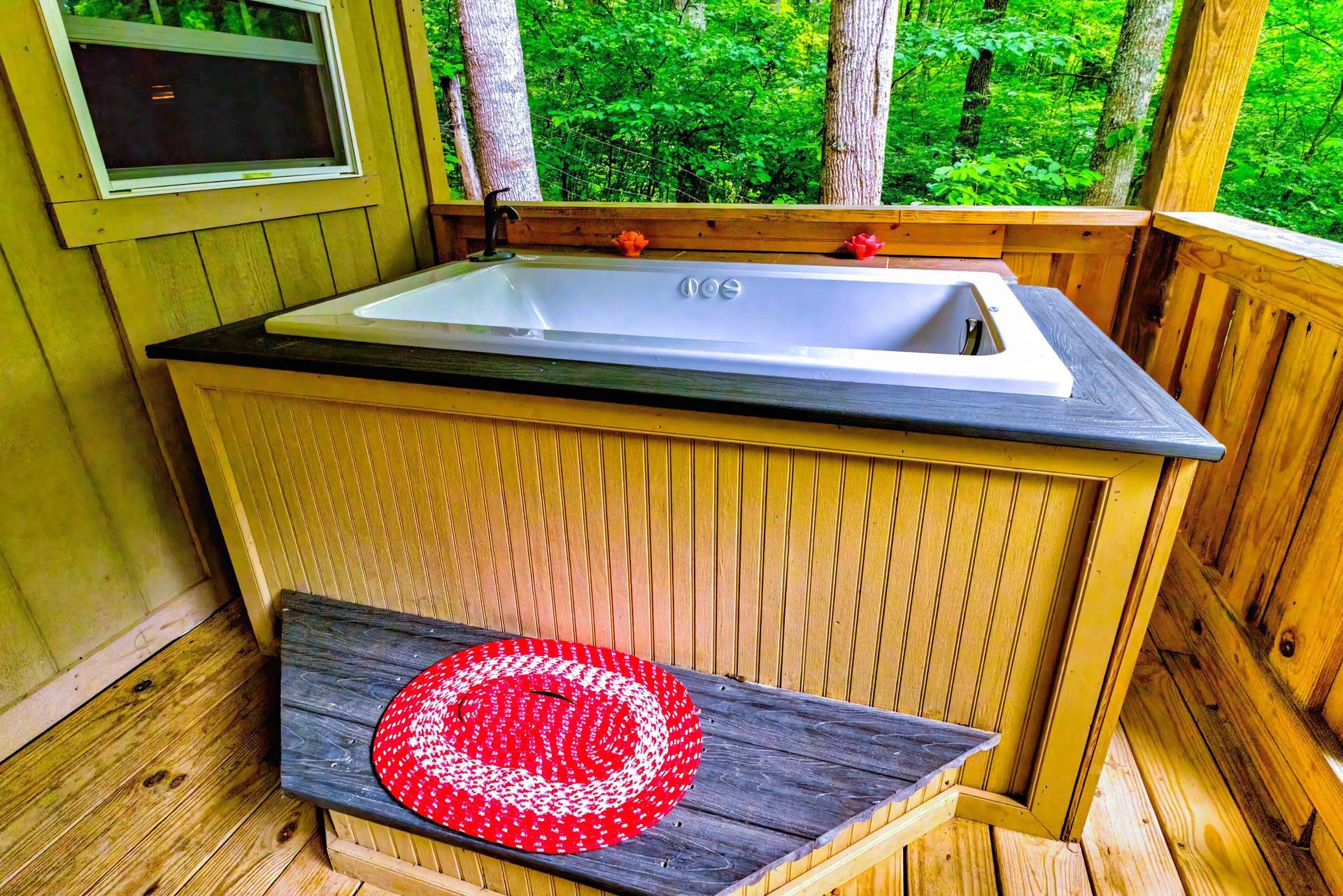 Outdoor hot tub on a wooden deck, with a red round cushion on a bench and green trees around.