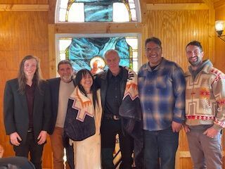 Six people standing together in a wood-paneled room with a stained-glass window backdrop