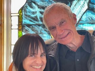 Two people smiling indoors in front of a colorful stained-glass window