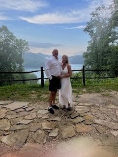 Couple standing on a stone overlook with trees and mountains under a cloudy sky.