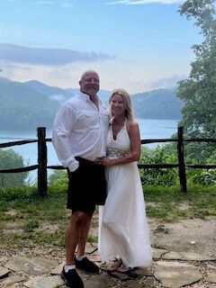 Couple in formal attire posing by a lakeside with mountains in the background