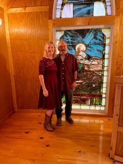 Two people standing in a wood-paneled room by a large stained-glass window.