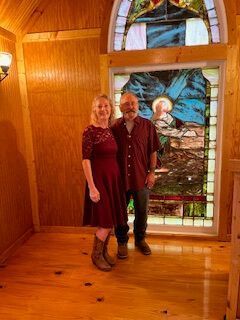 Two people stand in a wood-paneled room beside a stained-glass window.