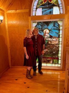 Two people standing in a wooden room beside a stained-glass window and door, with sunlight streaming in.
