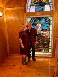 Two people standing in a wooden room beside a stained-glass window.