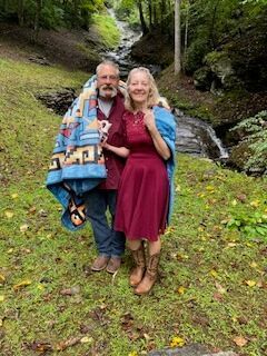 Two people smiling and standing close together in a grassy woodland by a small stream.