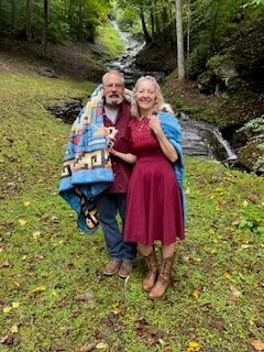 Couple standing in a wooded creekside clearing, wrapped in a colorful blanket and smiling at the camera.