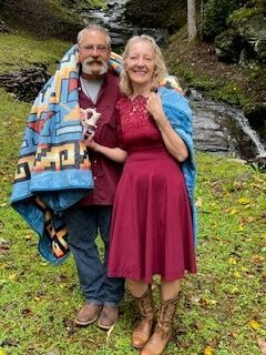 Smiling couple standing outdoors on grass, wrapped in a colorful blanket, near a small waterfall