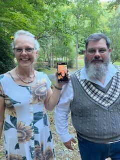 Two people outdoors in a wooded area, smiling while one holds up a phone displaying a portrait.