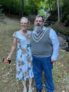 Two people smiling on a wooded trail, standing close together outdoors.