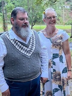 Two people standing outdoors, smiling in a wooded garden, one in a gray sweater vest and one in a white floral dress