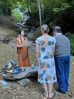 Woman speaking outdoors beside a small waterfall, facing two people in a forest clearing.