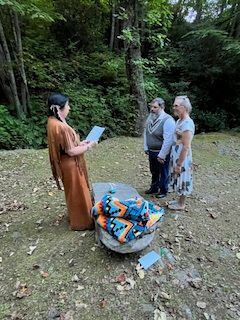 Three people stand in a forest clearing while one reads from a paper, with a colorful bundle on the ground.