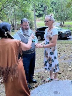 Three people standing outdoors in a wooded area, talking near parked cars