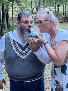 Two people outdoors share a drink from a small cup in a wooded area.