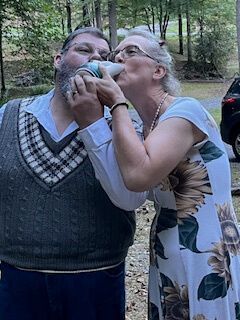 Two people outdoors sharing a drink from the same bottle, standing in a wooded area.
