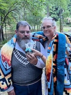 Two people wrapped in colorful blankets stand outdoors, smiling and holding a small plush toy.