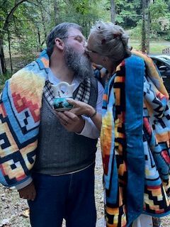 Two people wrapped in colorful blankets lean in for a kiss outdoors, one holding a drink.