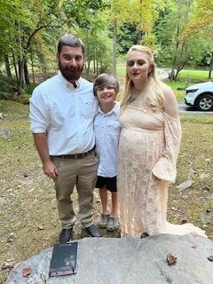 Three people posing outdoors near a gravestone in a wooded area
