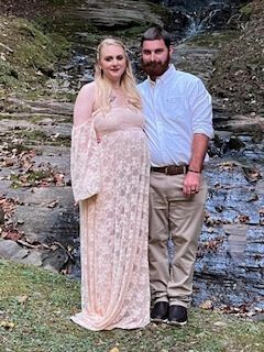 Couple standing on rocks by a stream, dressed in formal attire, with greenery in the background