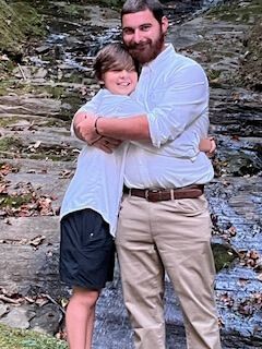 Two people hugging outdoors by a rocky stream, both in light shirts and khaki pants.