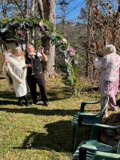 People posing under a flowered arch outdoors while a person holds a camera; lawn chairs in foreground.