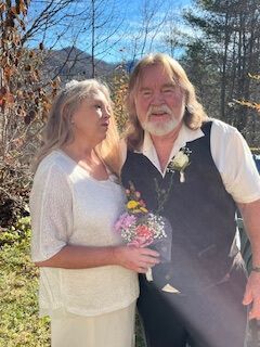 Couple posing outdoors in sunlight, holding a bouquet, with bare trees and a blue sky behind them
