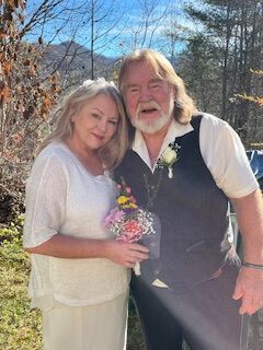 Couple posing outdoors, smiling and holding flowers in a sunlit wooded setting.