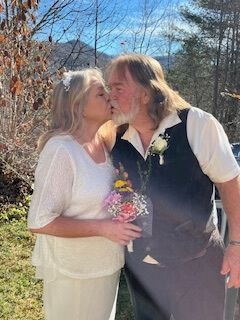 Two people kissing outdoors in sunlight, one holding a small bouquet, with trees in the background.