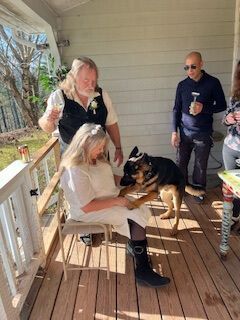 Two people and a dog on a porch, with one person sitting and petting the dog