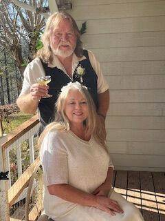 Two people on a porch, one seated and smiling, one standing behind holding a drink.