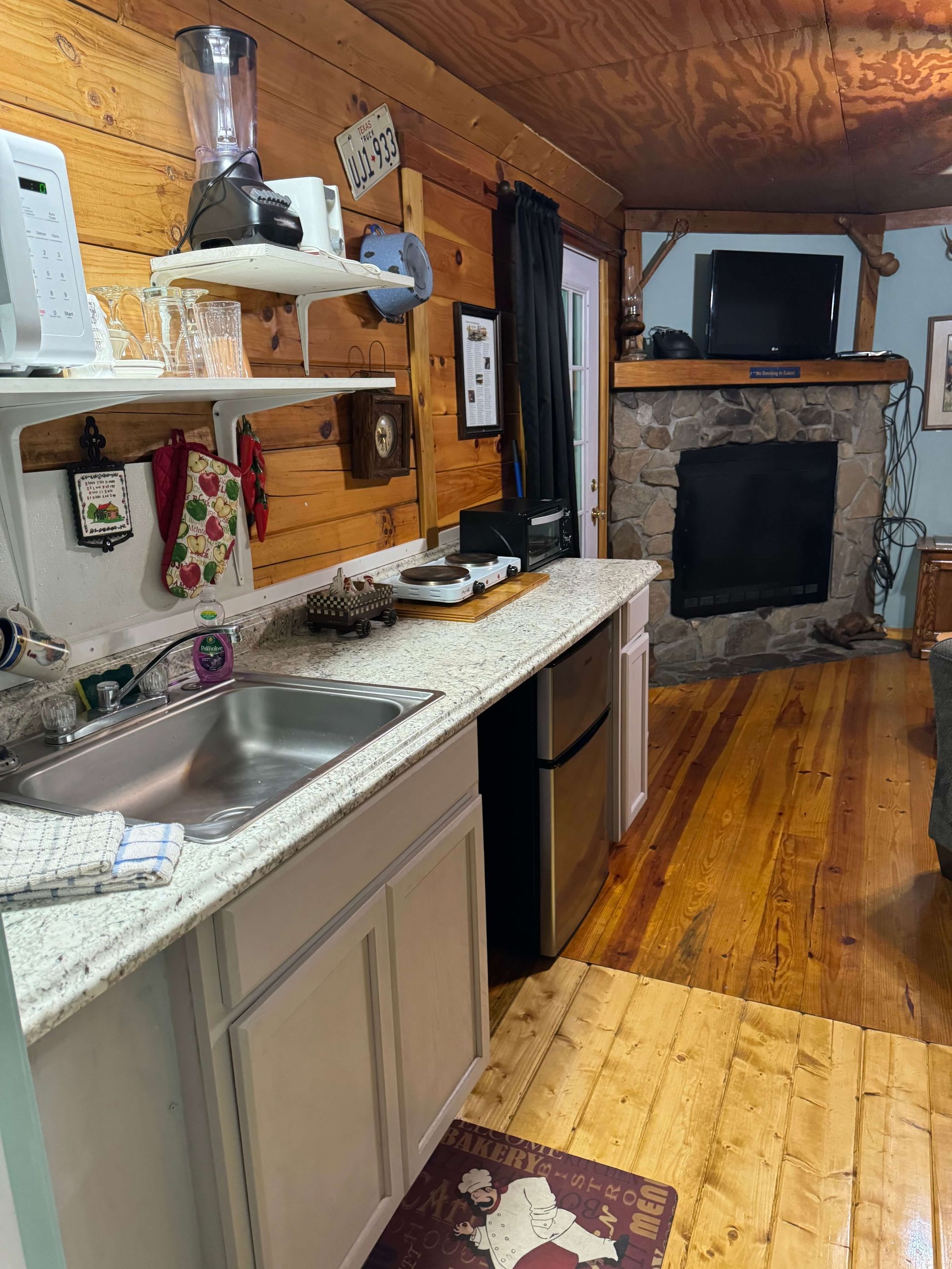 Rustic kitchen with white cabinets, speckled countertop, stone fireplace, and wood-paneled walls.