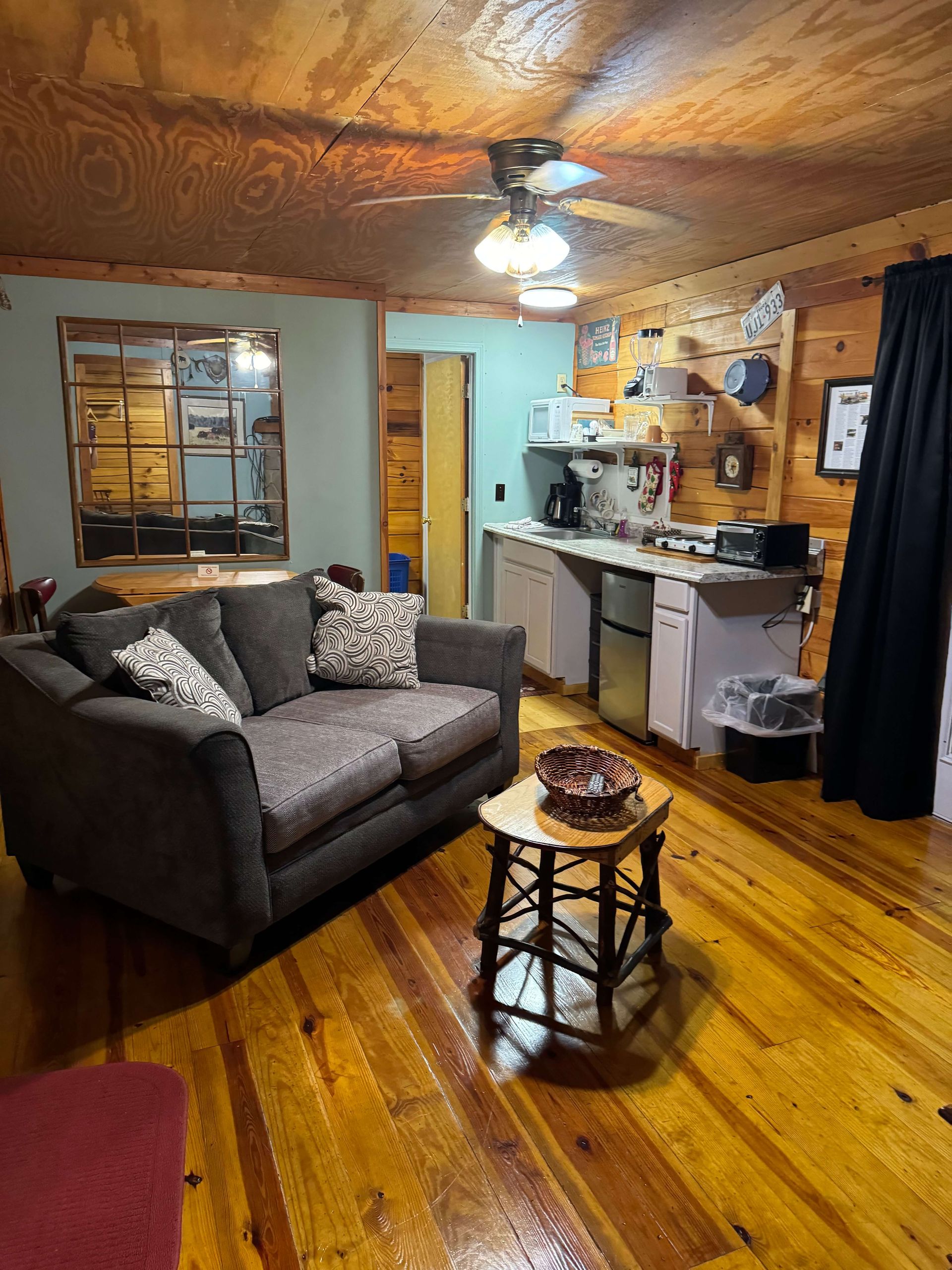Cozy living room with gray loveseat, wooden floor, ceiling fan, and kitchenette in the background