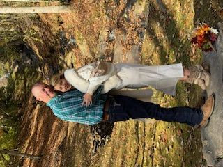 Couple in wedding attire standing outdoors beside a tree and flower bouquet