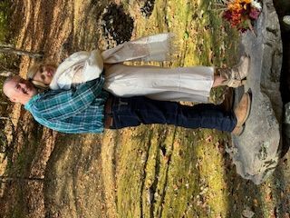 Two people standing outdoors on a rocky riverbank, with a waterfall in the background.