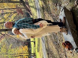 Two people standing outdoors in an autumn park beside a stream and rocks, with fallen leaves around them