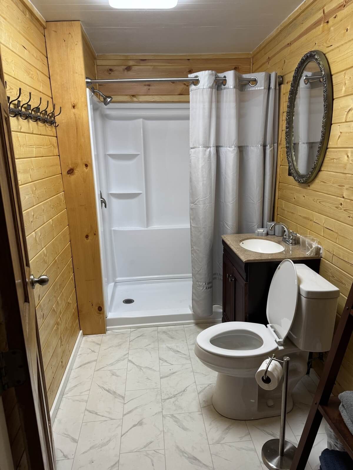 Narrow bathroom with shower, toilet, sink, wood-paneled walls, and a round mirror