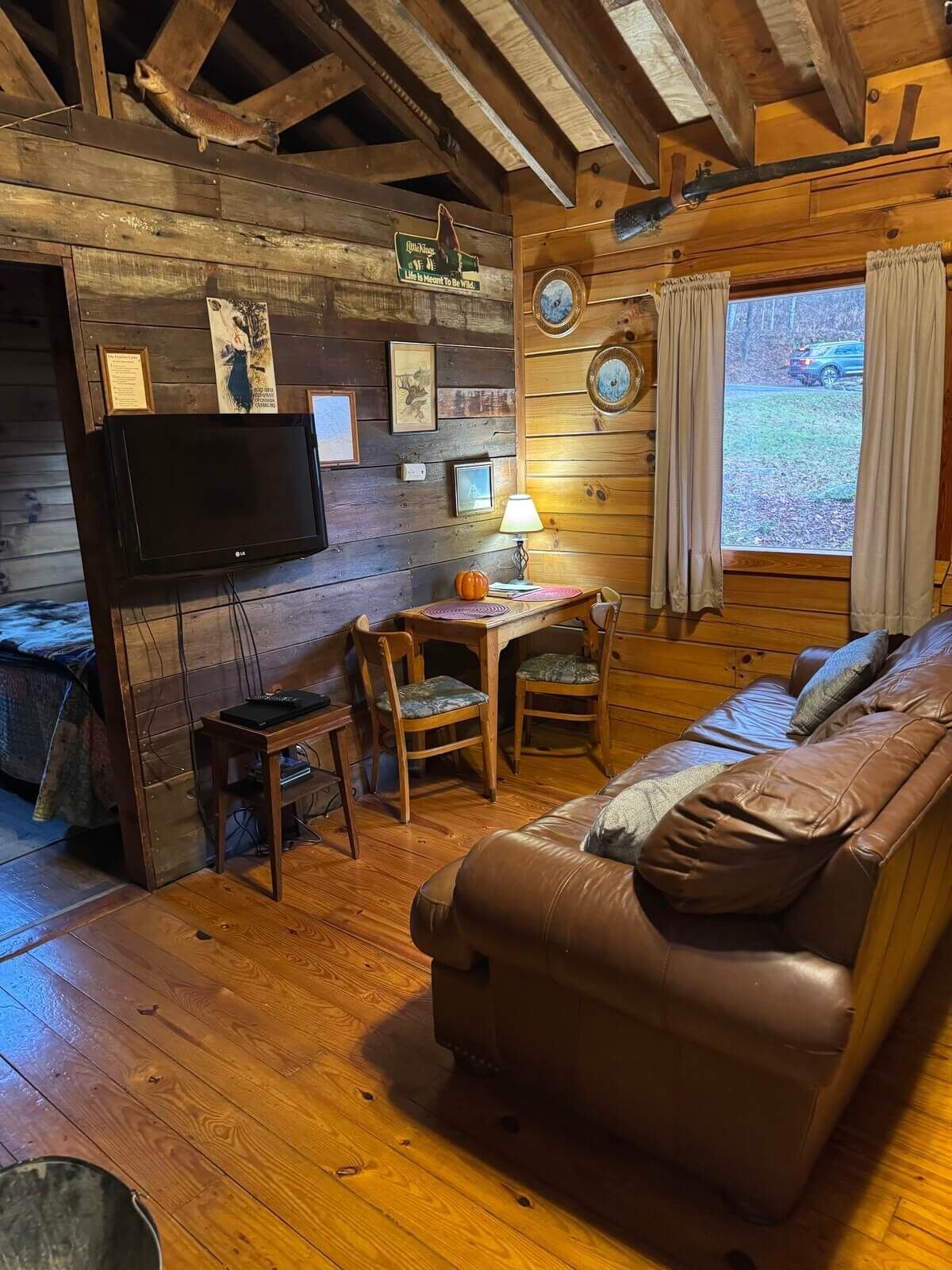 Cozy rustic cabin living room with leather sofa, wood stove, TV, small table, and a large window by the lake.