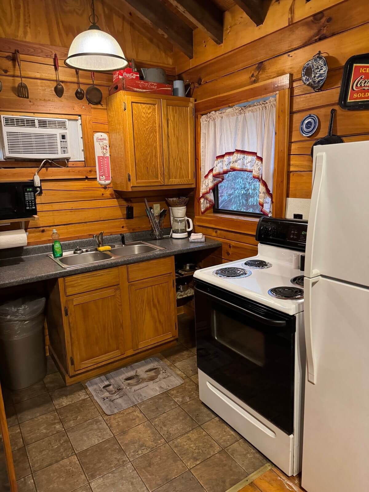 Small rustic kitchen with wood cabinets, white stove and fridge, and a sink under a curtained window.