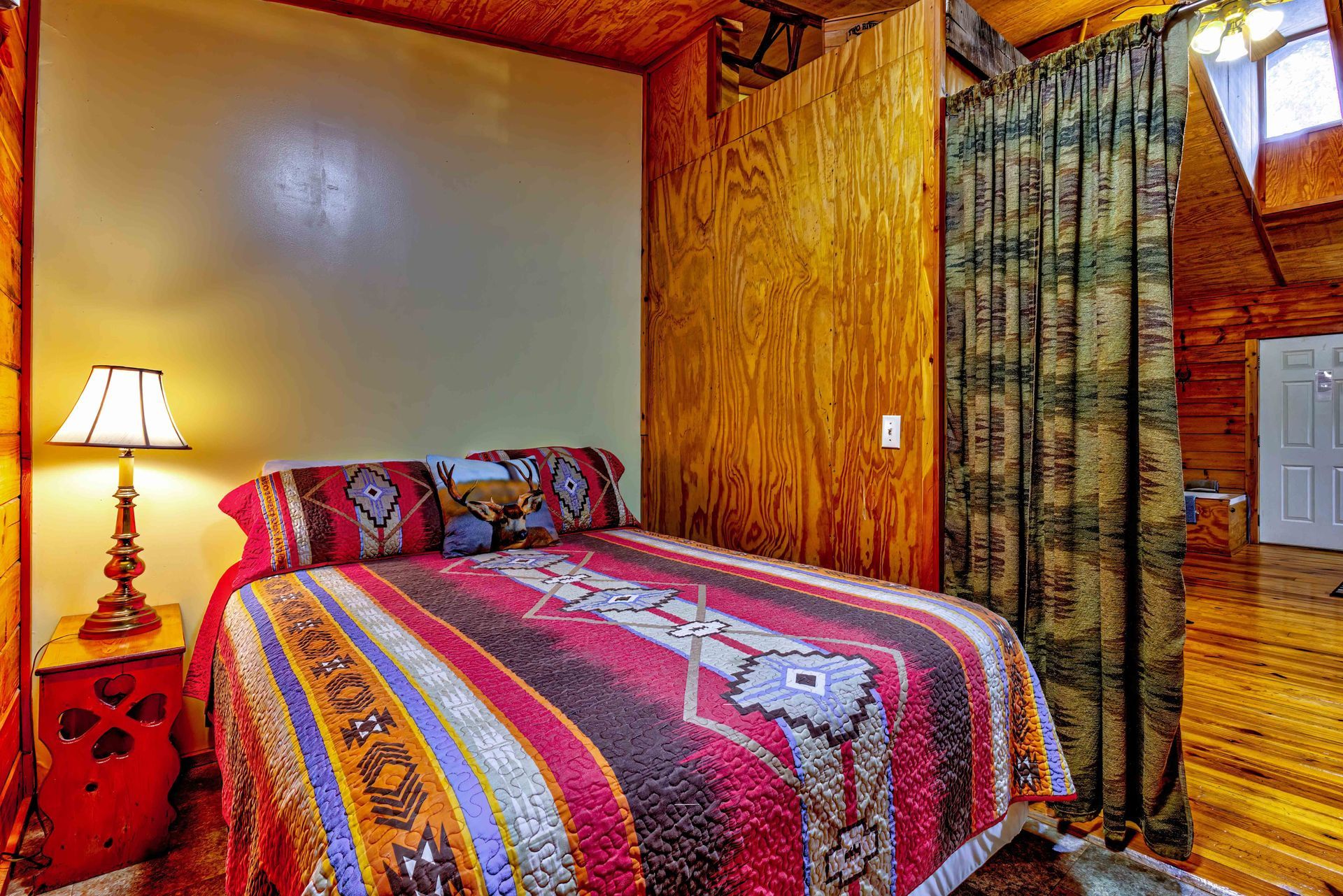 Cozy bedroom with a colorful patterned bedspread, bedside lamp, wood-paneled walls, and tiled floor.