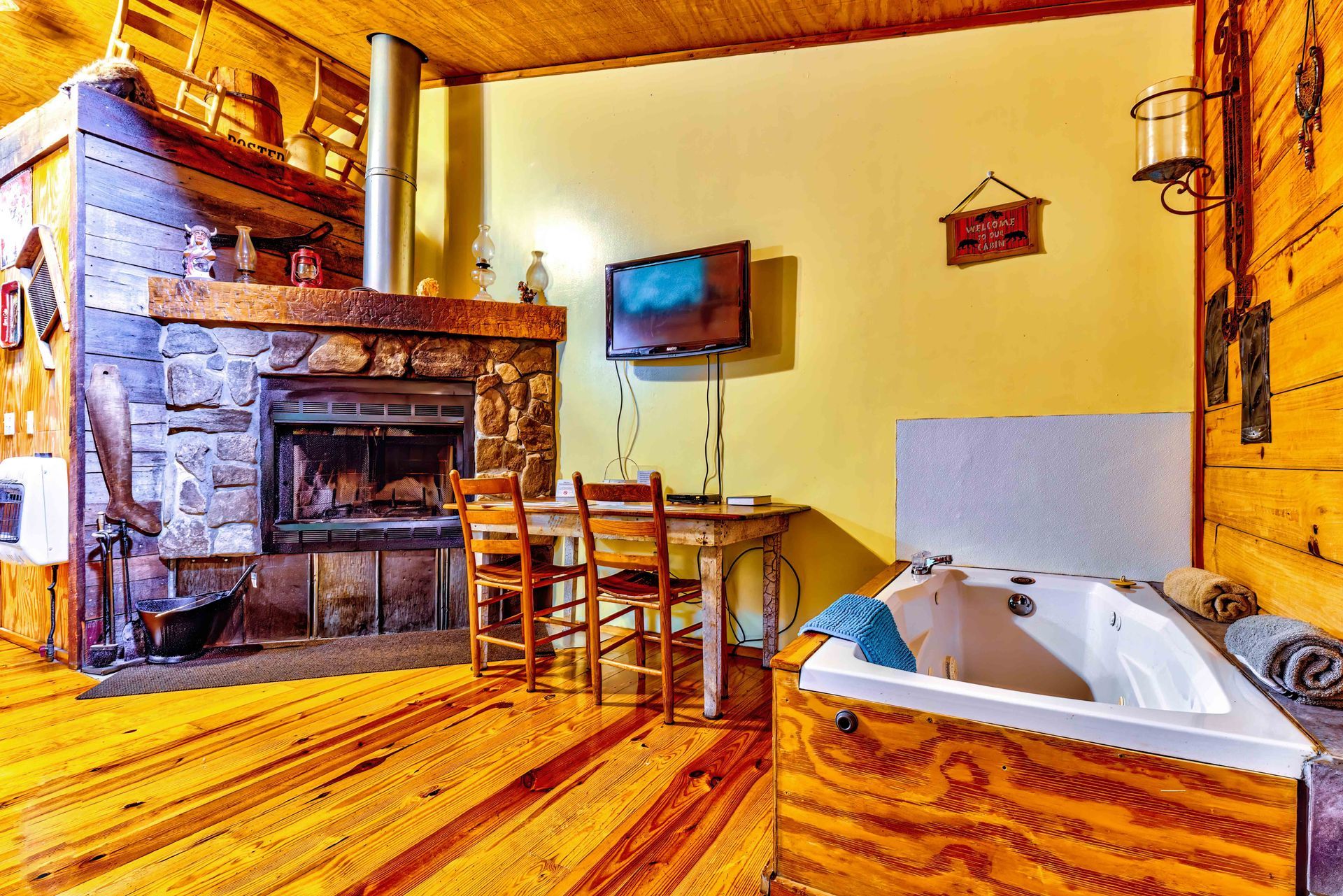 Cozy cabin bathroom with wood floors, stone fireplace, dining table, and a white corner jacuzzi tub