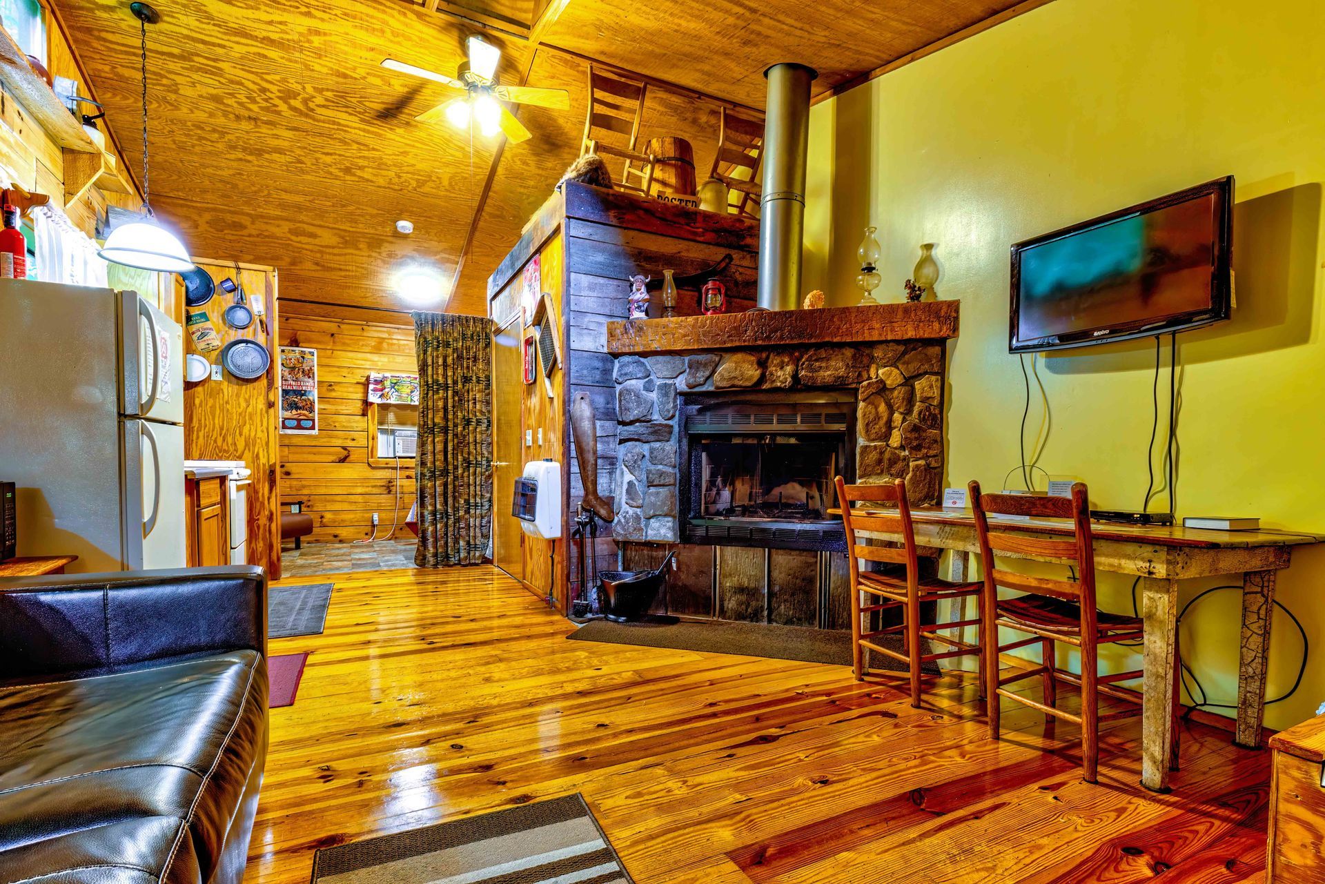 Cozy rustic cabin living room with wood floors, stone fireplace, dining table, and wall-mounted TV