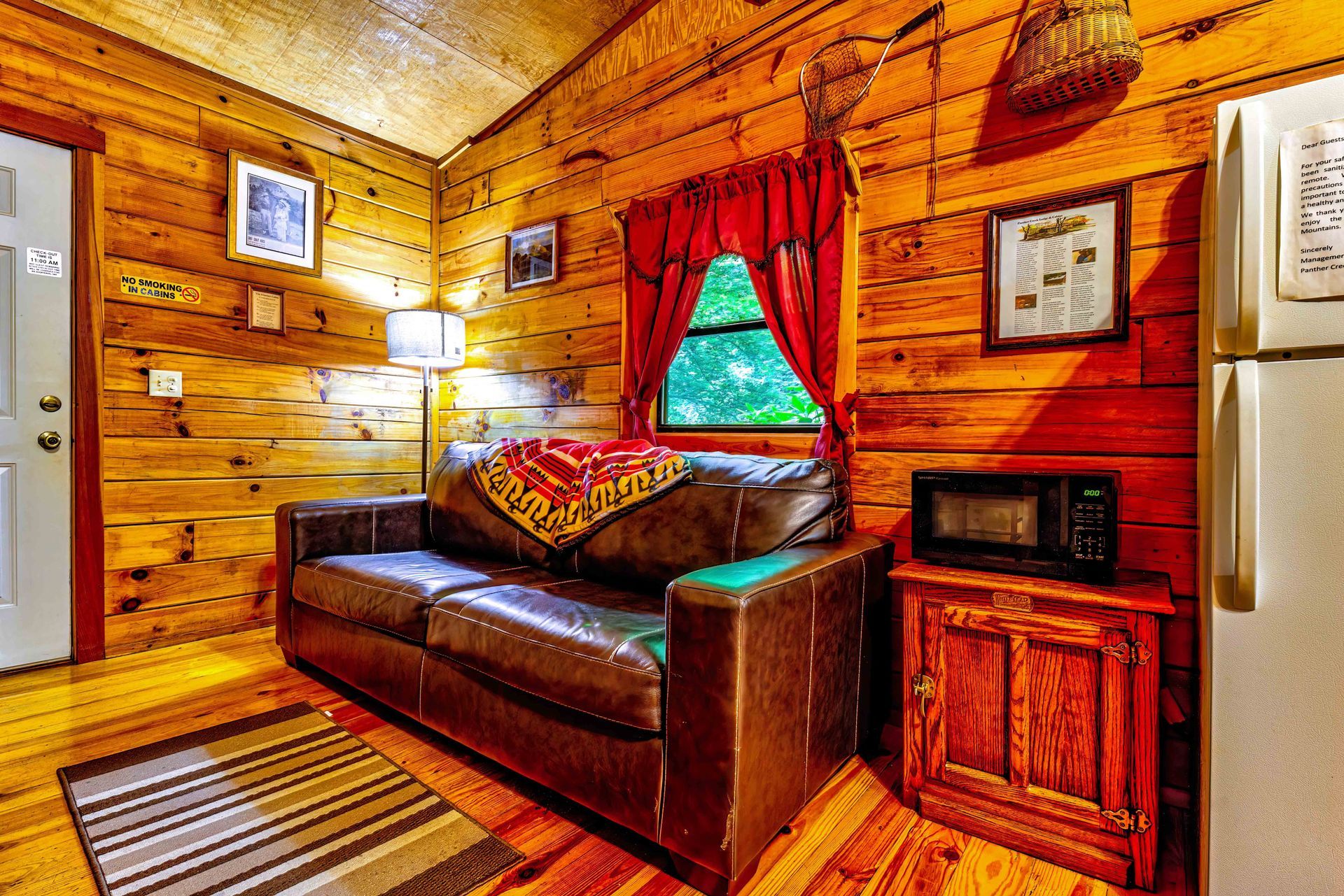 Cozy wood-paneled cabin bedroom with a leather sofa, red curtains, and a mini fridge and microwave.