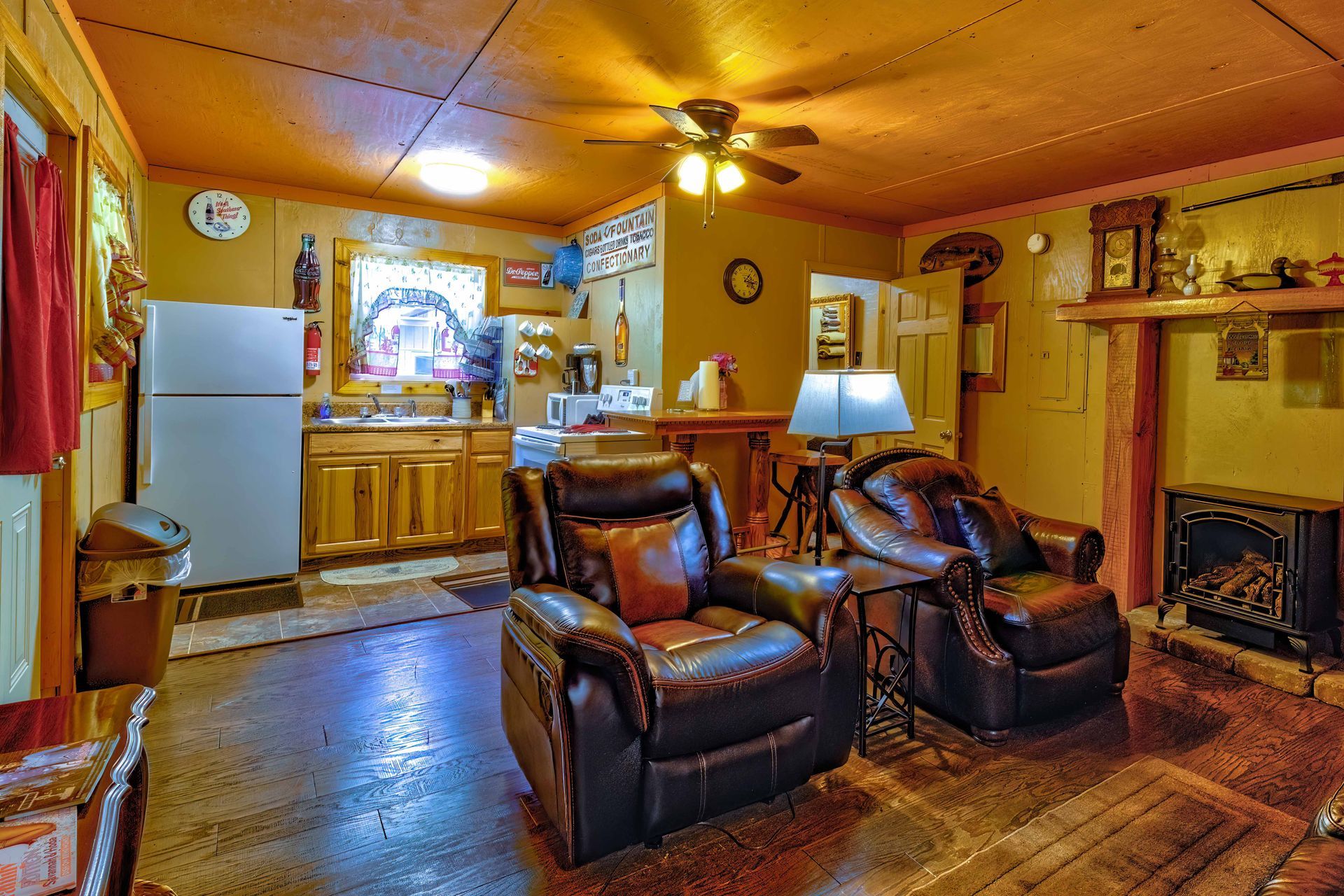 Warm living room with leather chairs, fireplace, wooden cabinets, and a kitchen area in the background