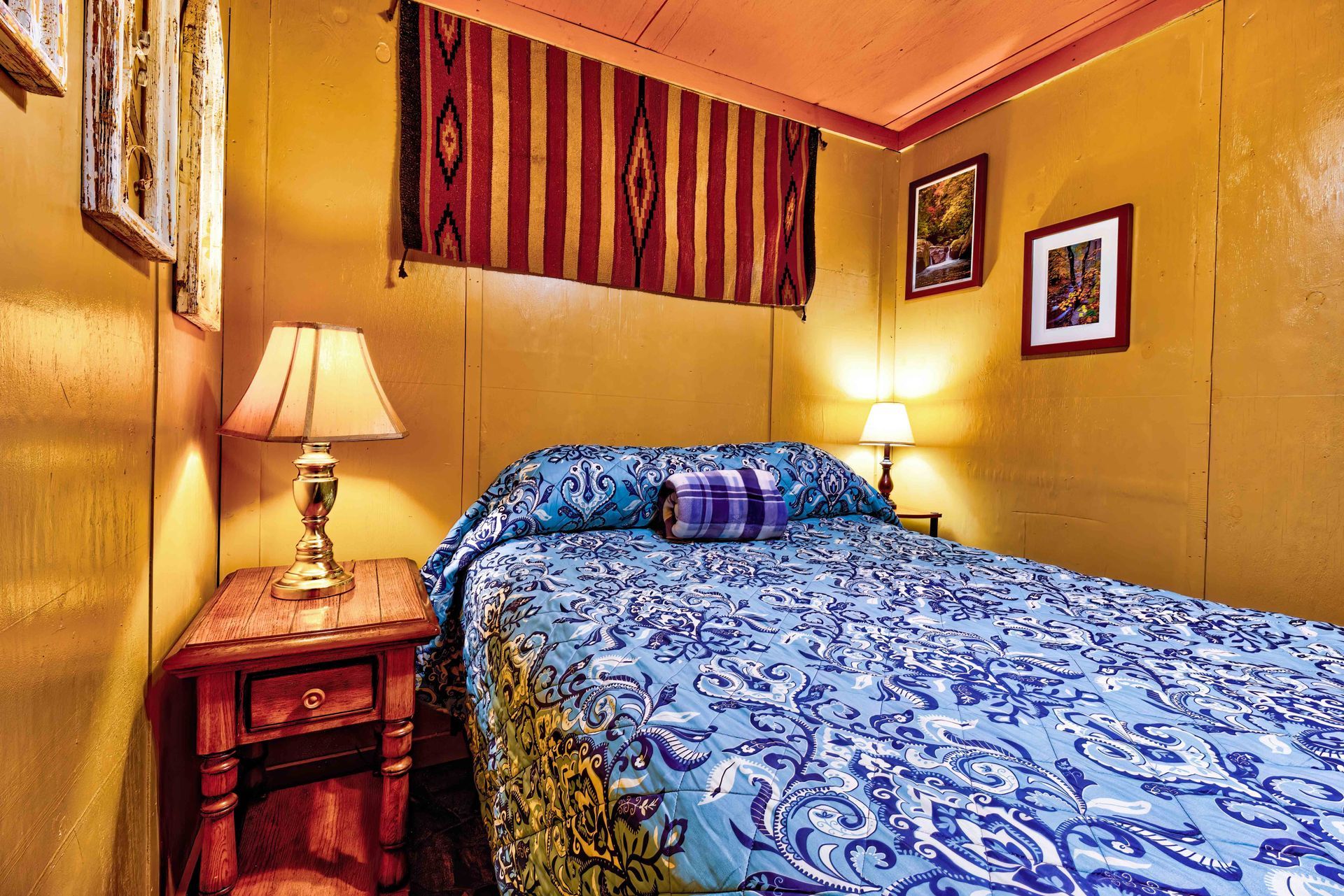 Small yellow bedroom with a blue patterned bedspread, two lamps, and framed wall art.