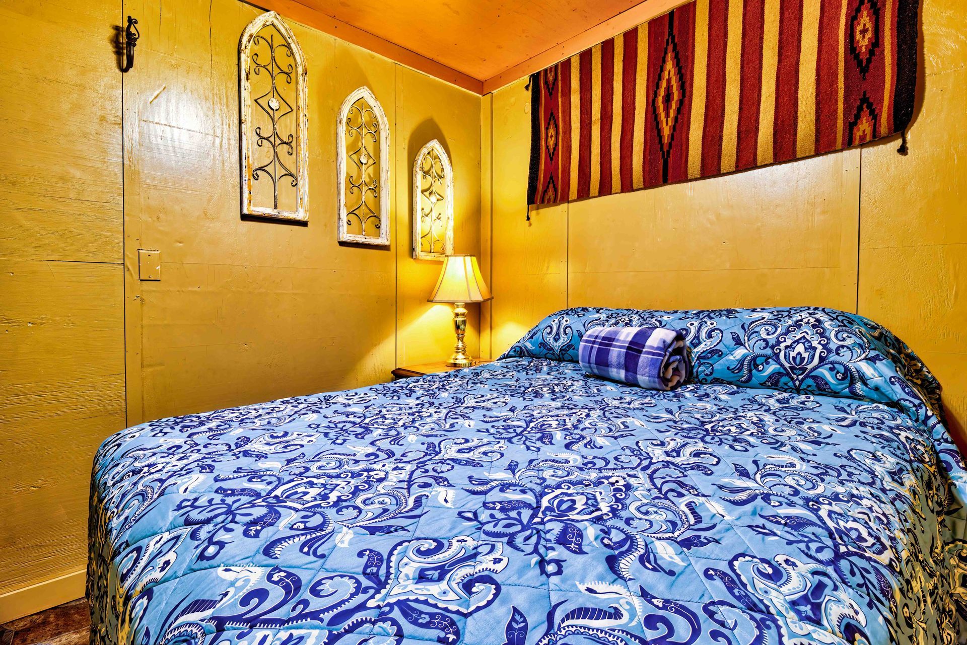 Cozy bedroom with blue patterned bedspread, warm yellow walls, lamp, and decorative wall hangings