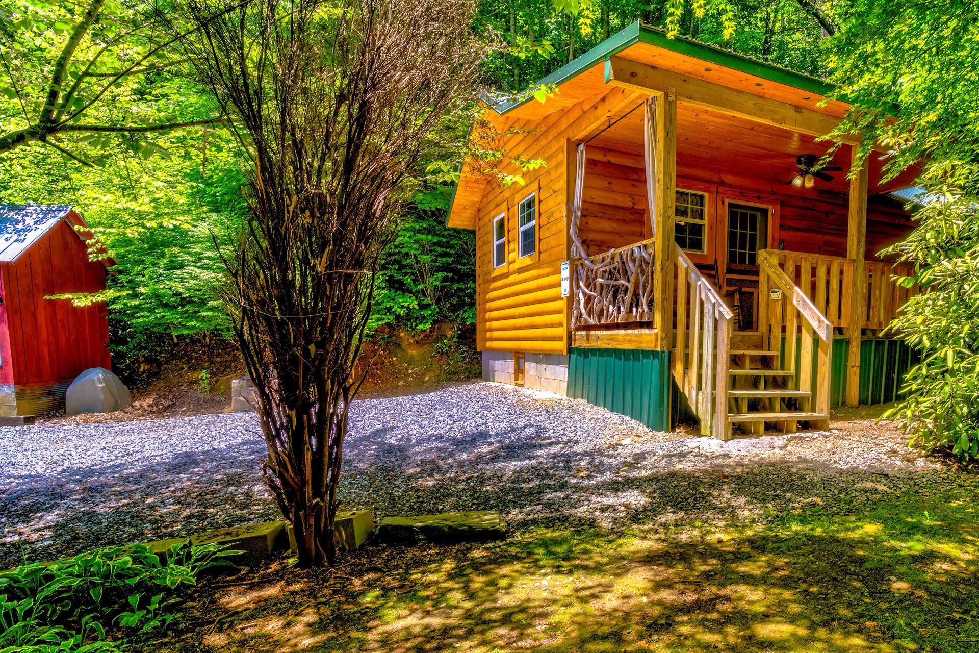Yellow cabin with a porch and stairs in a wooded yard, with a red shed nearby