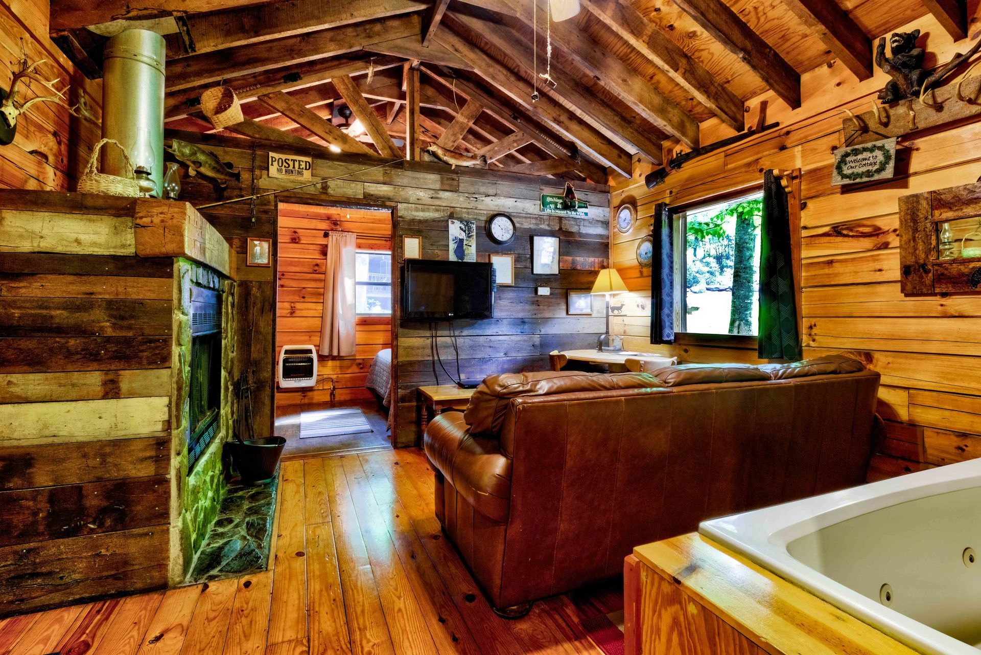 Rustic cabin living room with log walls, leather couch, wood stove, and a white bathtub in the foreground