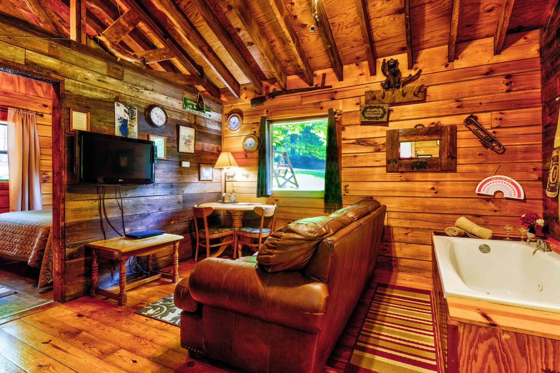 Rustic log cabin living room with wooden walls, TV, sofa, desk, and a window view of green trees