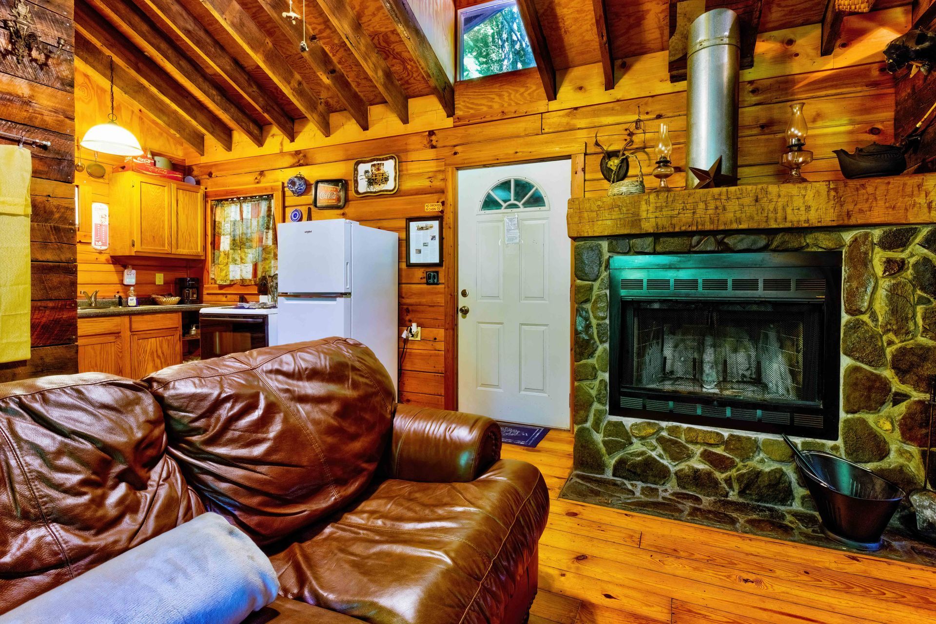 Cozy log cabin living room with leather sofa, stone fireplace, wood beams, and white refrigerator